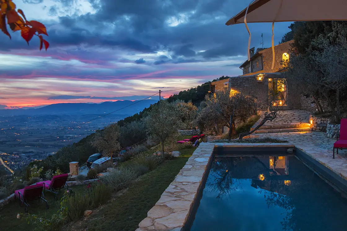 Perfect Hideaways real estate property: Warm yellow light shines from the Tuscan style villa. Sunset view from the natural stone patio beside the outdoor swimming pool. Casa Di Led in Spoleto.