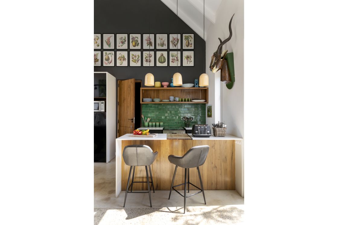 Modern kitchen with bar seating, overhead lighting, and green tiled backsplash above the stove. Grey walls decorated with vintage botanical prints. Overberg.