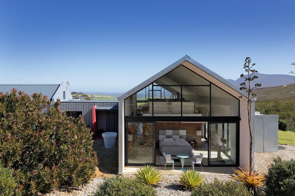 An A frame-house nestled among lush trees and green grass, with a backdrop of mountains.