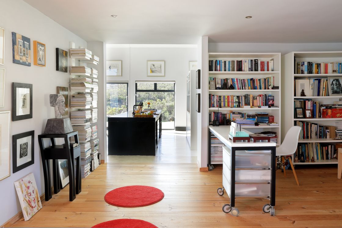 A cozy library and study room featuring a desk, bookshelves, and a rug, seamlessly connecting to an open-plan kitchen.