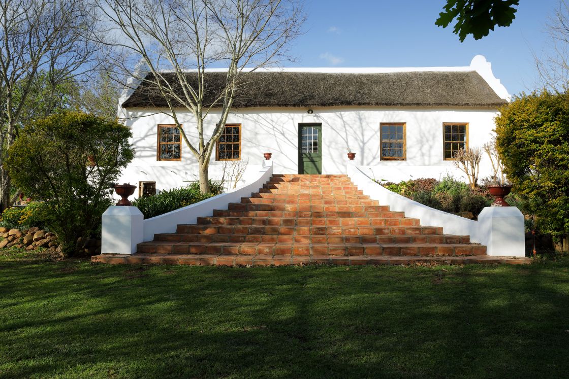 Outdoor steps leading to thatch-roofed Cape-Dutch farmhouse. Central green door and symmetrical window placement.