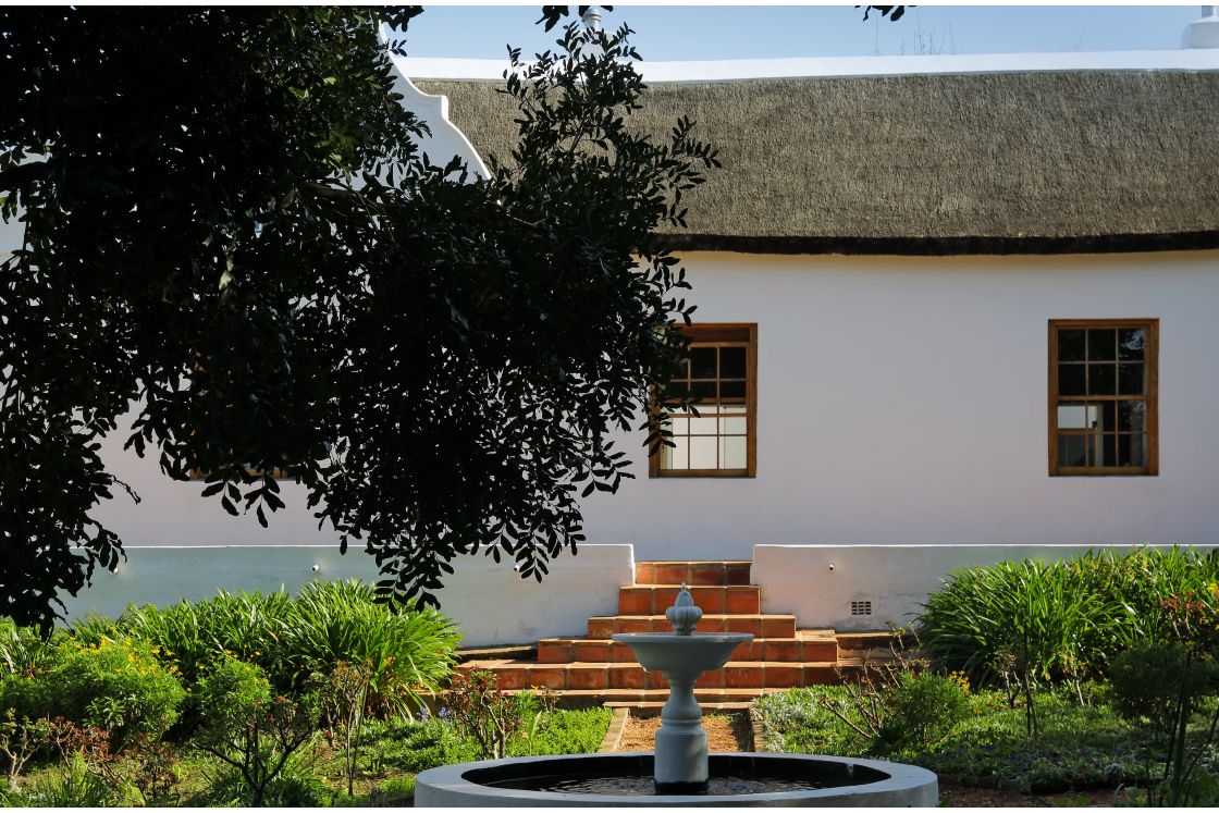 Garden courtyard water feature surrounded by symmetric footpaths, a nearby Cape-Dutch style homestead with connected patio steps.