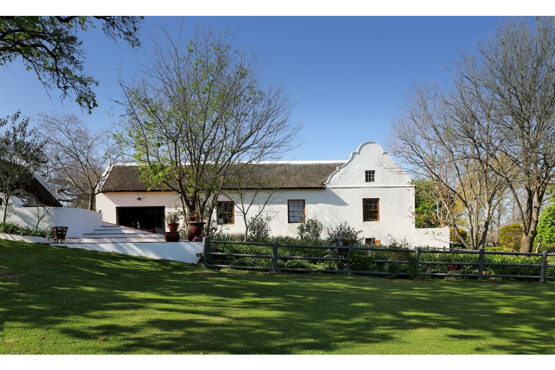 Landscape view of main homestead with grass and simple wooden fence. Steps lead towards the patio and outdoor fireplace.