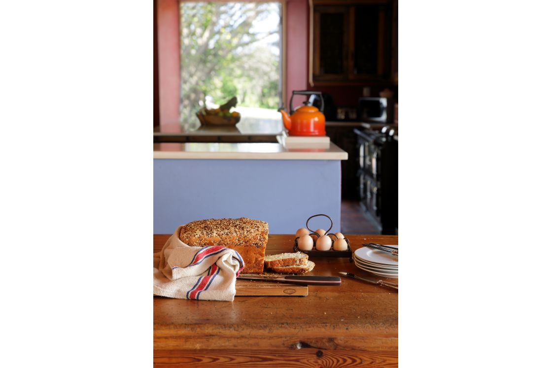 Farm style bread, eggs, and cutlery on a wooden table in the foreground. Blue kitchen island with kettle and garden view in the background.