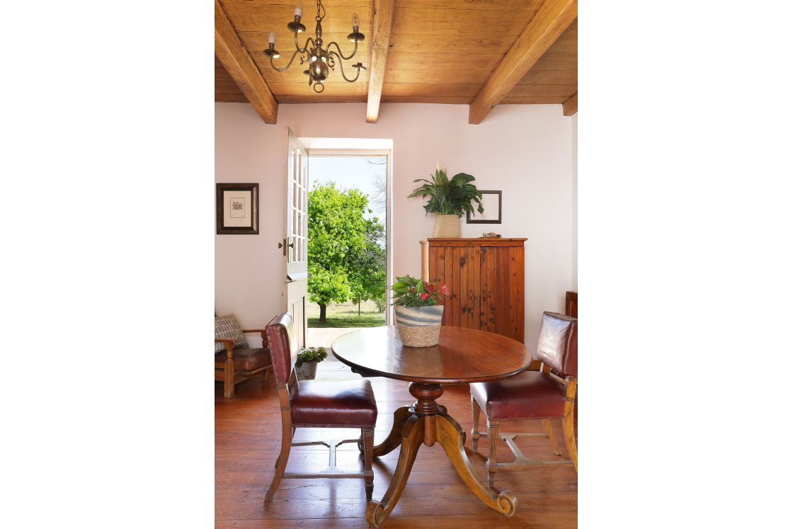 Interior view of farmhouse, two leather chairs set around ornate dining table, wooden cabinet against back wall, with garden view through back door.