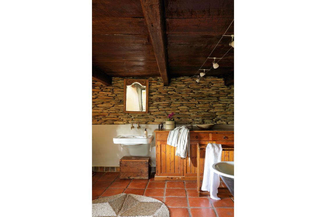 Heritage-style bathroom featuring raw stone walls and dark ceilings. Basin, free-standing bath, and wooden cupboard.