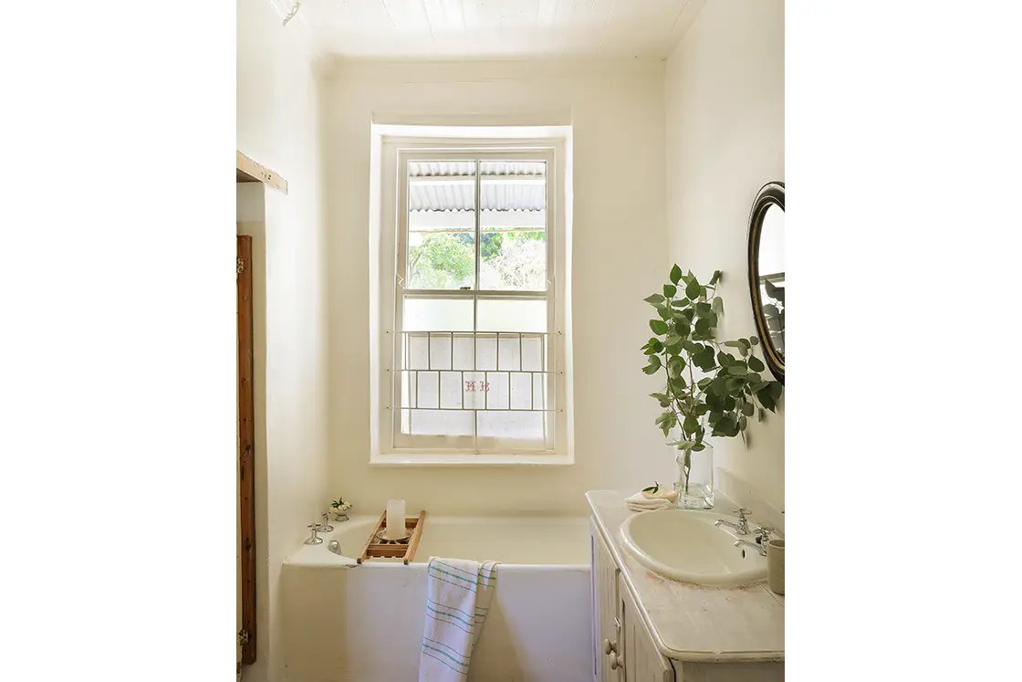 Perfect Hideaways real estate property: Bathroom with corner bath, wooden bathroom cabinet, basin, and a mirror. Clothes and decorative branches placed on the wall hanger. The White House in Barrydale.