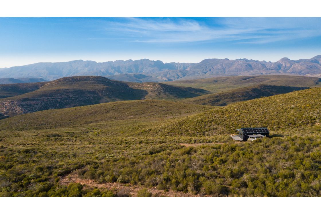 A homestead on a largely undeveloped plot of land, aside from the dirt road leading to the home, expansive mountain ranges fill the background.