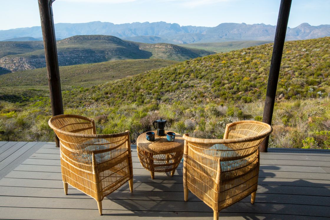 A wicker coffee table and chairs on the shaded patio of a small homestead, with views of the mountainous terrain.