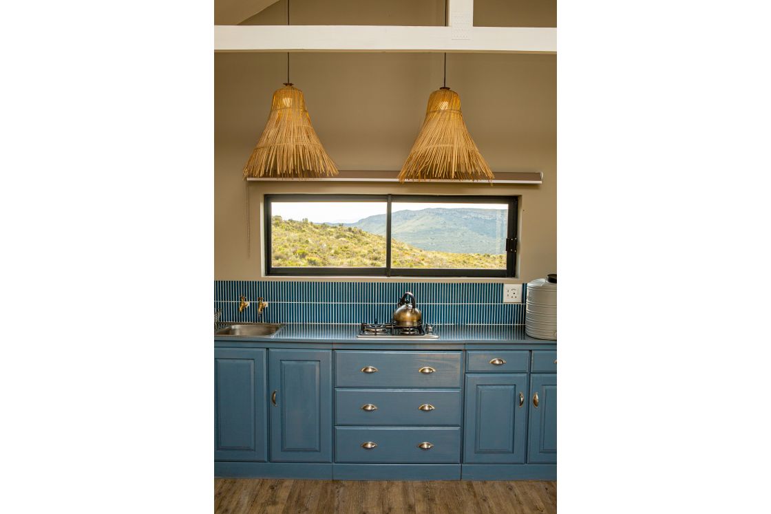 A kitchen area with blue cupboards and backsplash, featuring a gas stove, sink unit, woven lighting fixtures, and views of the surrounding mountain ranges.