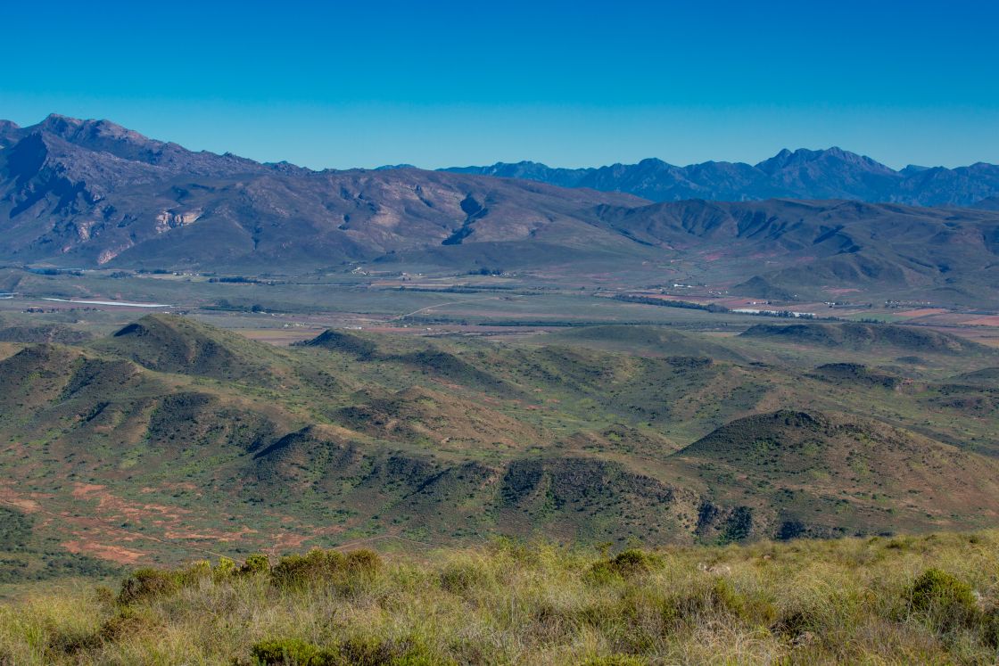 Undulating hills give way to a farmland area at the foot of an expansive mountain range.