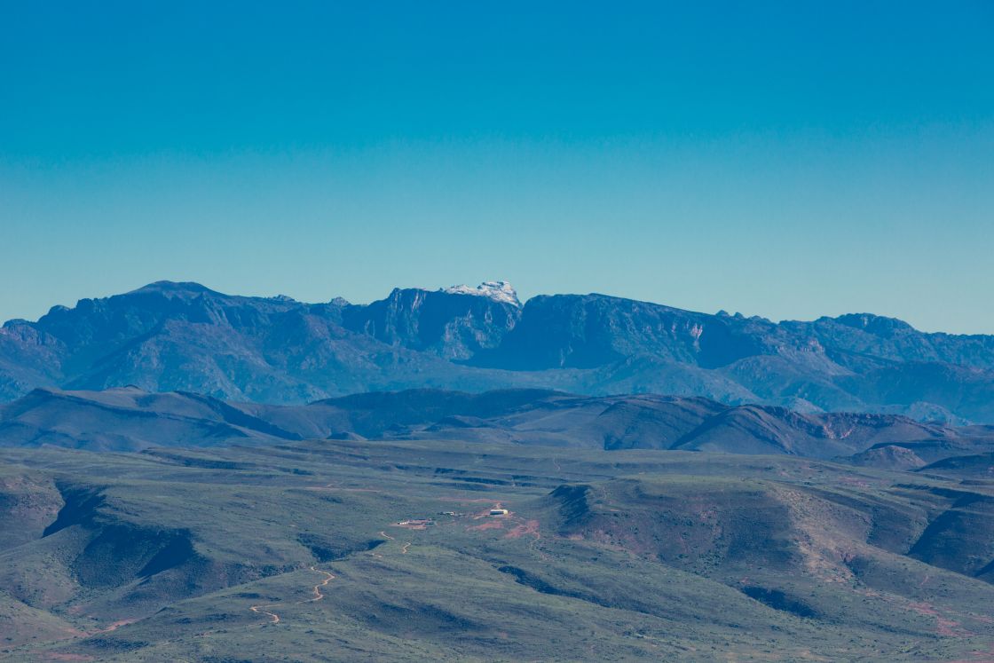 A landscape view of the undulating Overberg hills, featuring a plot of land with a dirt road leading to the homestead, and a snow-capped mountain in the center of a broad range of mountains.