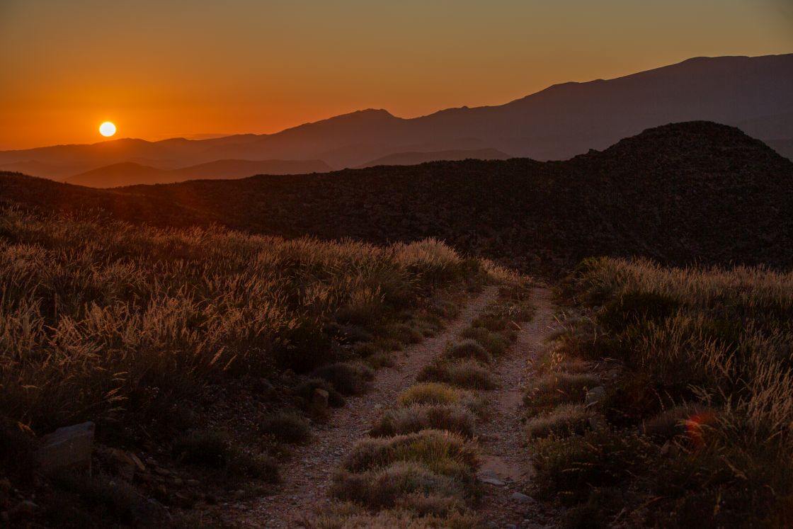 The sunset casts an orange glow over the grassy vegetation which borders a dirt road, undulating hills and misty mountain ranges fill the background.